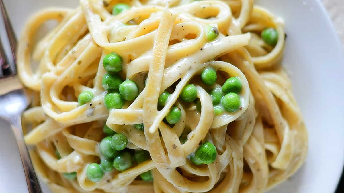 Close-up of a dish of creamy fettuccine pasta with green peas on a white plate. A silver fork is partially visible on the left side of the plate. The pasta is evenly coated with a rich, white Alfredo sauce, showcasing one of the finest Alfredo recipes, and the peas are nestled within the noodles.