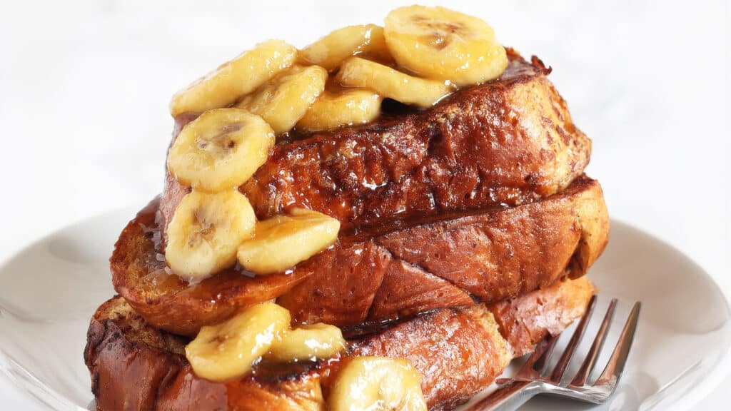 A plate with two thick slices of French toast stacked on top of each other. The toast is covered with syrup and topped with sliced bananas. A fork lies next to the toast on the white plate, reminiscent of your favorite pancake recipes for a touch of morning nostalgia.