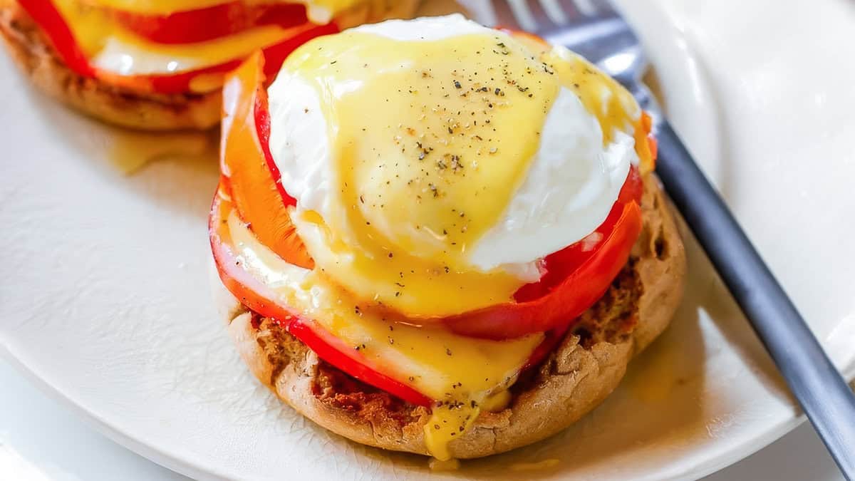 A close-up of a dish featuring a toasted English muffin topped with a slice of tomato, a poached egg, and hollandaise sauce. The sauce is garnished with ground black pepper. Perfect for breakfast or light lunch and dinner, this delightful creation is paired with a fork on a white plate.