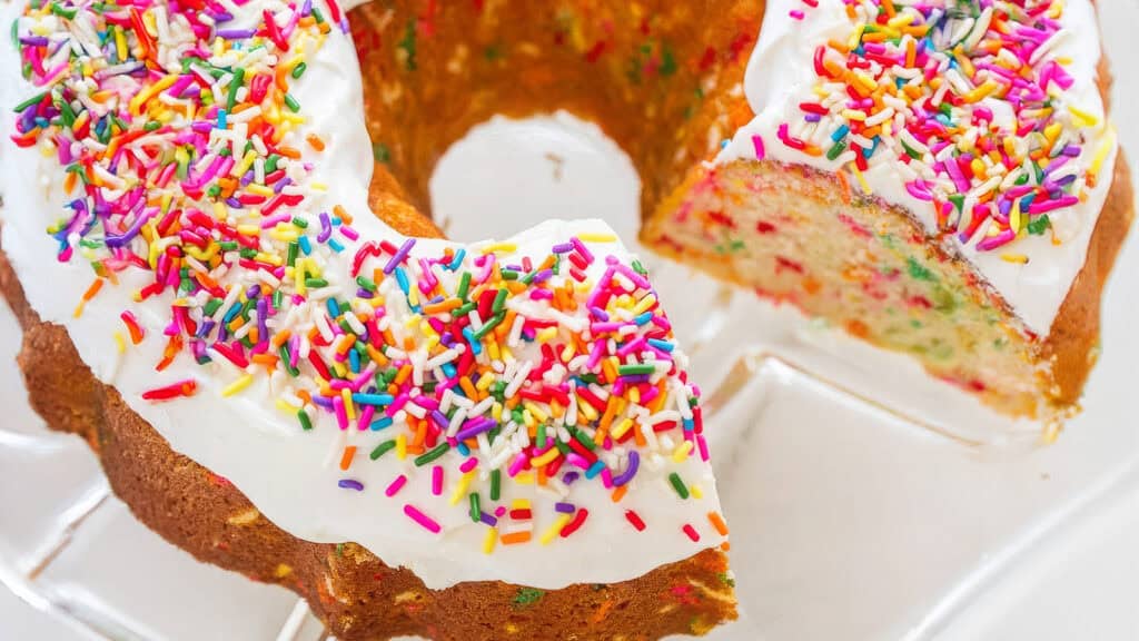 A round, colorful funfetti bundt cake topped with a white glaze and multicolored sprinkles sits on a clear cake stand. A slice has been cut and partially removed, revealing additional rainbow sprinkles baked into the cake—perfect for fans of unique Bundt Cake Recipes.