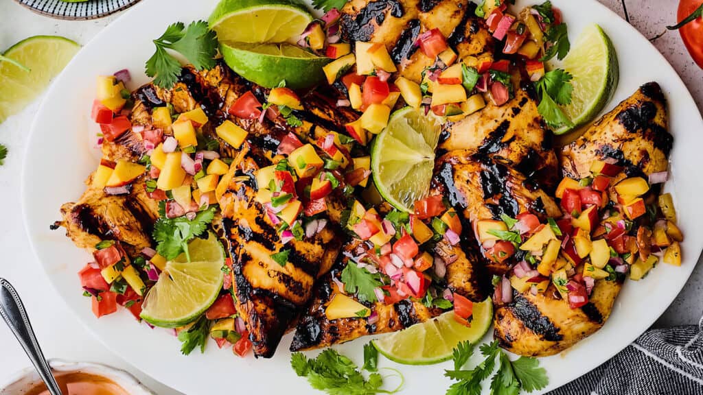 Grilled chicken recipes topped with a colorful mango salsa, garnished with lime wedges and cilantro, served on a white platter. The salsa includes diced mango, tomato, red onion, and herbs, creating a vibrant and appetizing dish.