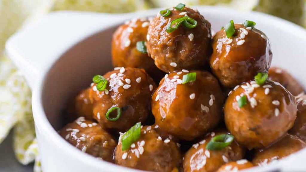 A close-up shot of a bowl filled with glazed meatballs, featuring a delicious ground beef recipe. The meatballs are garnished with sesame seeds and chopped green onions, arranged neatly, with the glaze giving them a shiny appearance.