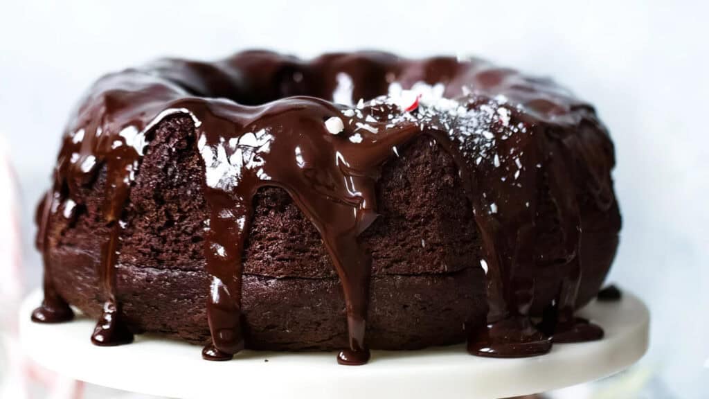 A rich, moist chocolate bundt cake displayed on a white cake stand. The cake is generously drizzled with glossy chocolate ganache, allowing it to cascade down the sides. The top is lightly sprinkled with powdered sugar, adding a festive touch. Perfect for those who adore Bundt Cake recipes!