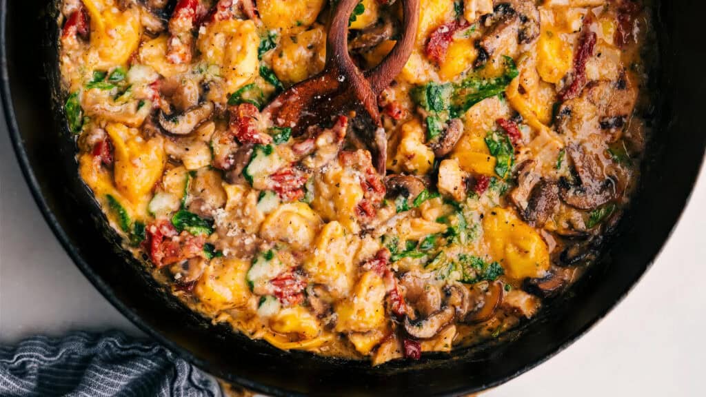 A skillet filled with creamy, stuffed pasta tortellini mixed with mushrooms, spinach, and sun-dried tomatoes, garnished with grated cheese. The dish has rich, vibrant colors that will wow pasta lovers. A wooden spoon rests in the pan, and a striped napkin is partially visible to the side.