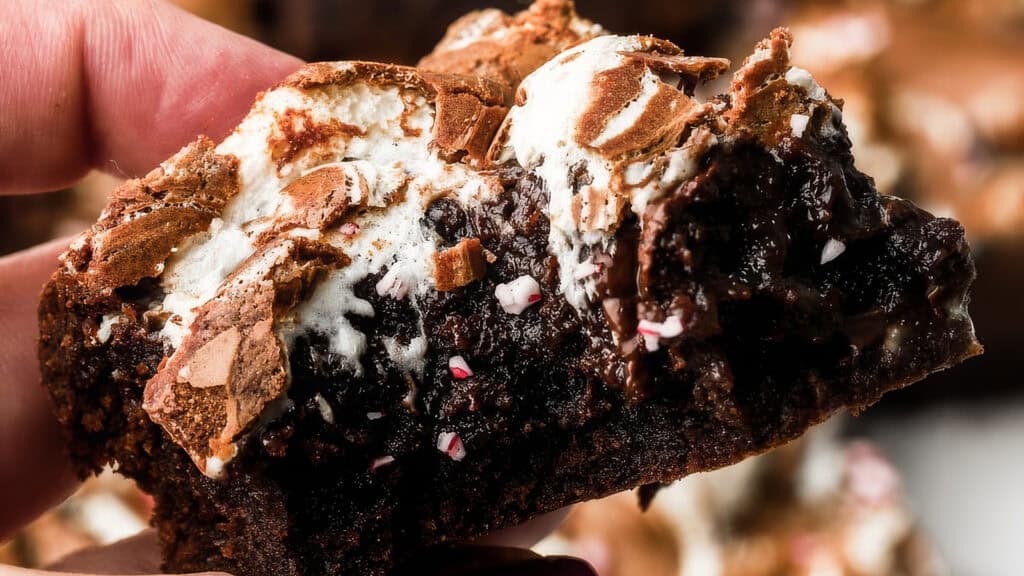 A close-up of a hand holding a gooey brownie topped with melted marshmallow and crushed peppermint pieces. Perfect for chocoholics, this brownie boasts a rich, moist texture with visible chocolate chunks. The slightly toasted marshmallow adds a crispy, golden-brown touch that goes beyond basic.