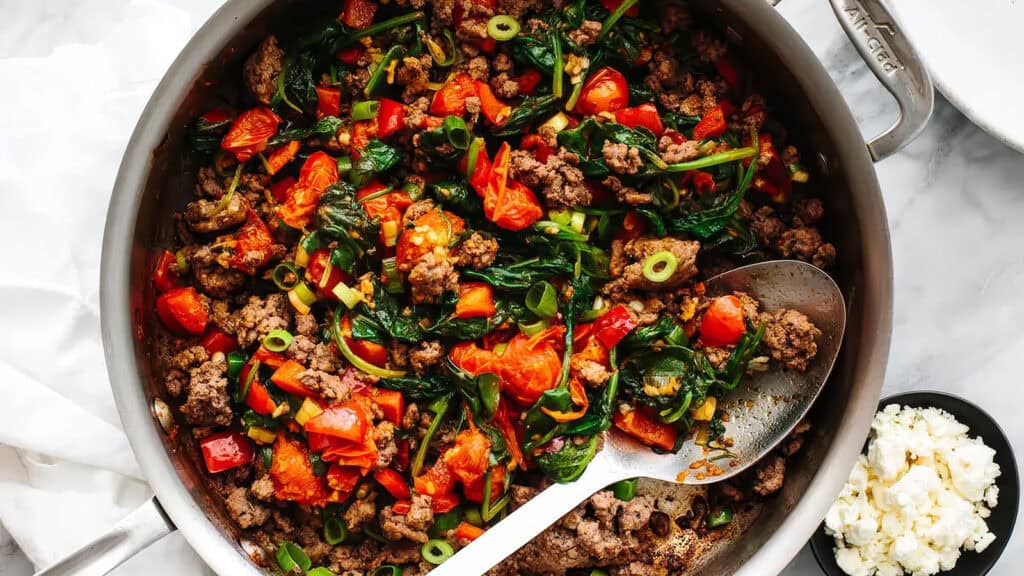 A pan filled with a colorful, cooked mixture of ground beef, spinach, chopped tomatoes, sliced green onions, and red bell peppers, with a spoon resting on the side. A small bowl of crumbled cheese is visible to the right of the pan. Perfect for those exploring new ground beef recipes.