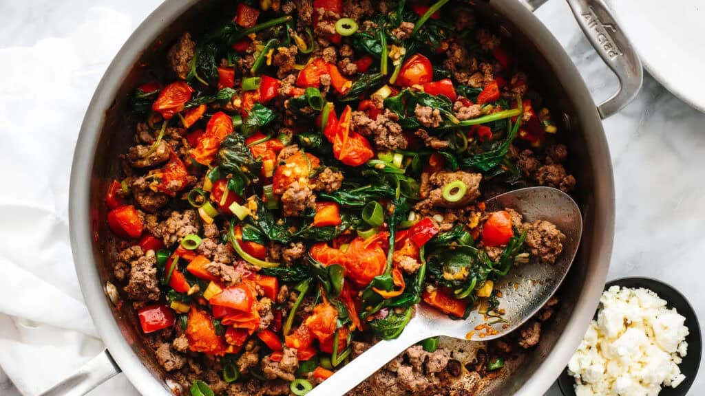 A skillet filled with a colorful sauté of ground beef, chopped tomatoes, spinach, diced bell peppers, and sliced green onions. A metal serving spoon rests inside the skillet. A small bowl of crumbled cheese is partially visible beside the skillet—a perfect addition to your ground beef recipes collection.