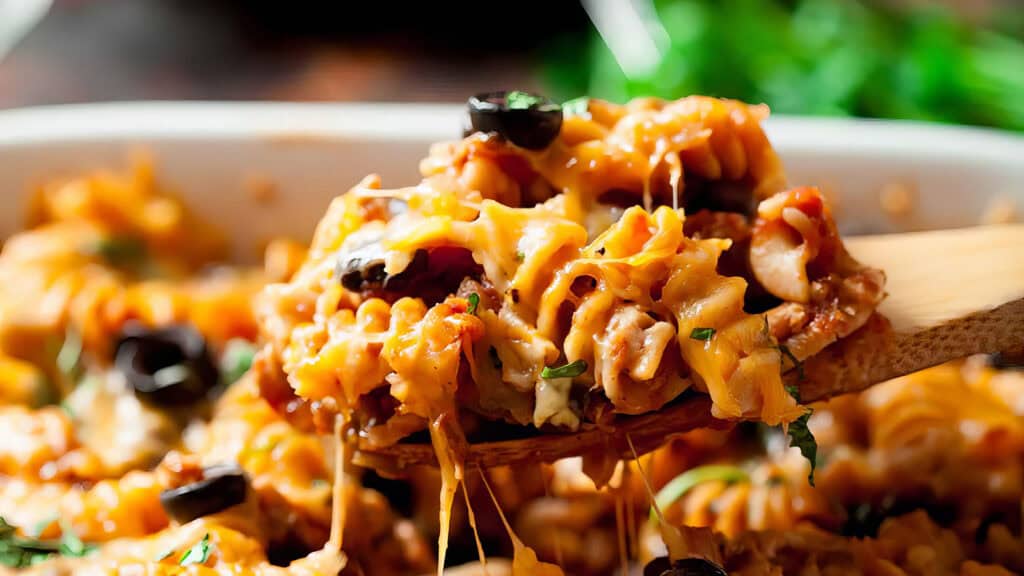 A close-up of a wooden spoon lifting a serving of cheesy baked pasta from a dish. This delightful casserole recipe features pasta topped with melted cheese, herbs, and black olives. The dish is garnished with fresh herbs, and green foliage is visible in the blurred background.