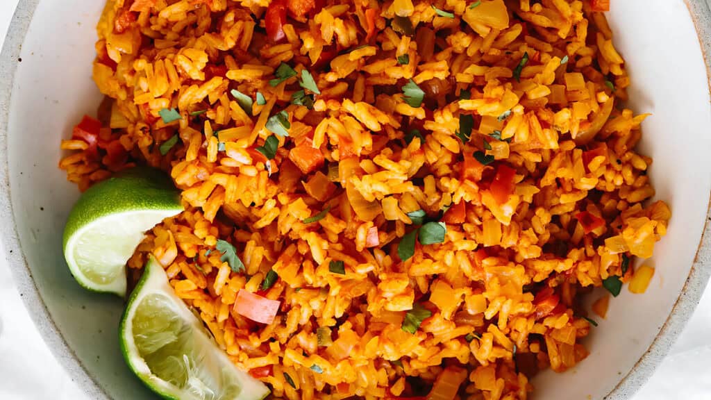 A close-up shot of a bowl of vibrant Mexican rice garnished with freshly chopped cilantro. The rice is mixed with finely diced red and green bell peppers and onions, a staple in many rice dish recipes. Two lime wedges are placed on the side.