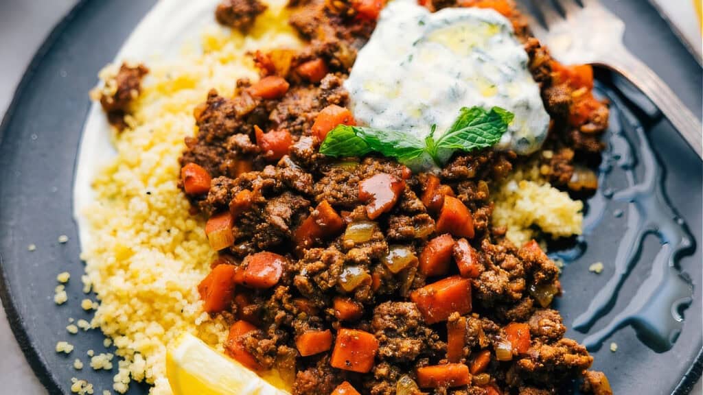 A plate of seasoned ground beef mixed with diced vegetables, served on couscous. The dish is garnished with a dollop of creamy sauce topped with herbs and a lemon wedge on the side. A fork rests on the plate. This is one of those hearty ground beef recipes that brings comfort to your table.