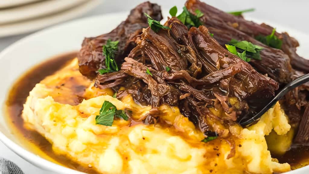 A close-up of a savory dish featuring tender, slow-cooked beef served over creamy mashed potatoes. The beef is garnished with fresh parsley and is accompanied by a rich, flavorful gravy that seeps into the mashed potatoes - perfect for those who love slow cooker recipes.