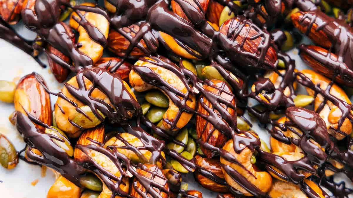 A close-up of a mix of almonds, cashews, and pumpkin seeds drizzled with dark chocolate. The nuts and seeds are laid out on a surface, and the dark chocolate is artistically poured over, creating an appetizing snack that will delight your taste buds. Perfect for fun treats or easy snacks!