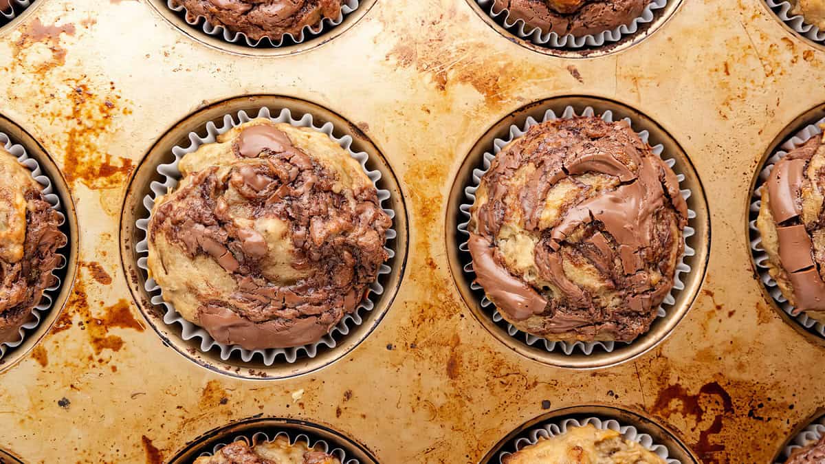 A close-up view of a muffin tray filled with freshly baked muffins swirled with Nutella. Each muffin is in a paper liner, and the tray shows signs of baking residue. The muffins have a marbled appearance from the chocolate spread mixed into the batter, perfect for your muffin recipes collection.