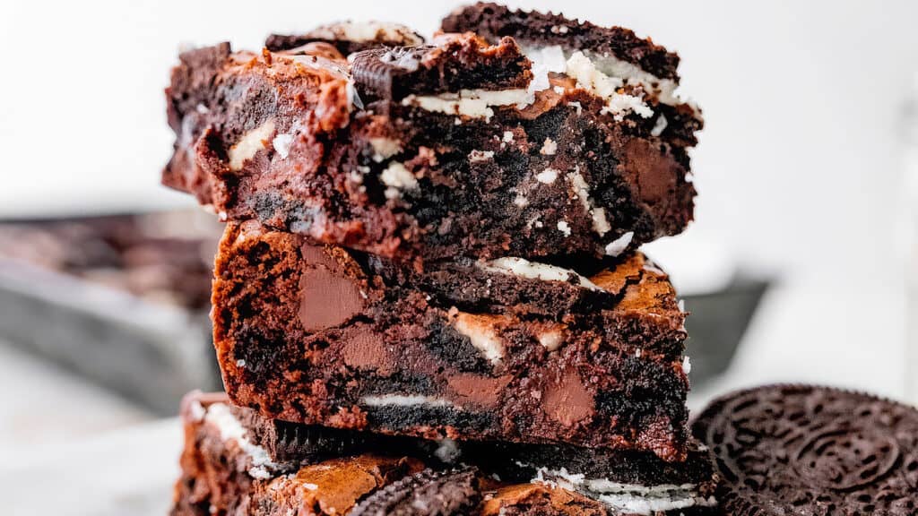 A close-up of a stack of brownies mixed with chunks of chocolate and Oreo cookies. The top brownie piece has a slightly crisp edge with a gooey, rich center. Crumbled cookies and chocolate pieces are visible throughout the dessert, making it look decadent and mouthwatering—a must-try for chocoholics seeking to go beyond basic brownie recipes.