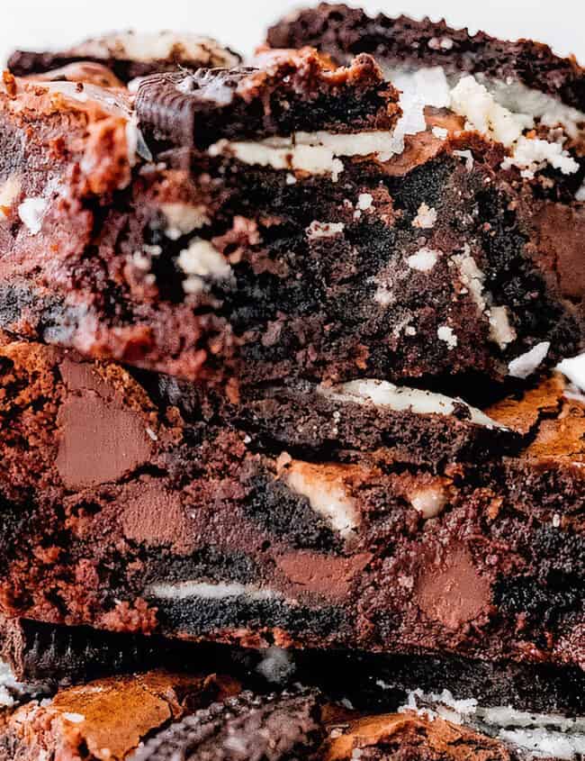 A close-up of a stack of brownies mixed with chunks of chocolate and Oreo cookies. The top brownie piece has a slightly crisp edge with a gooey, rich center. Crumbled cookies and chocolate pieces are visible throughout the dessert, making it look decadent and mouthwatering—a must-try for chocoholics seeking to go beyond basic brownie recipes.