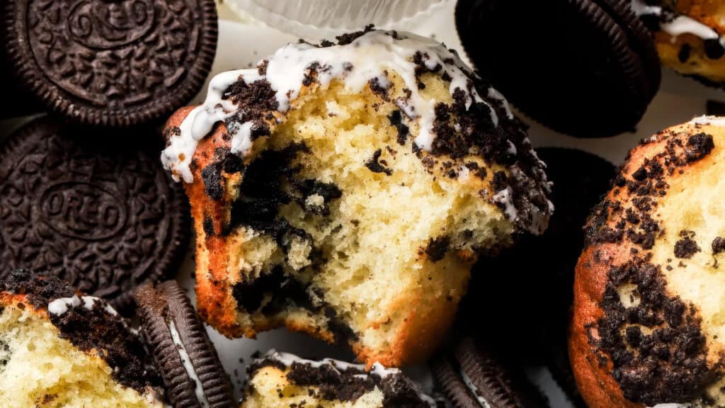 Close-up of a partially eaten muffin topped with crushed cookies and white icing, surrounded by whole sandwich cookies. The muffin has visible cookie pieces embedded in its fluffy interior, creating a visually rich contrast with the dark cookies around it—a delightful addition to your favorite muffin recipes.