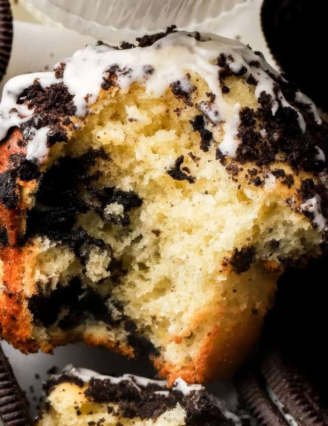 Close-up of a partially eaten muffin topped with crushed cookies and white icing, surrounded by whole sandwich cookies. The muffin has visible cookie pieces embedded in its fluffy interior, creating a visually rich contrast with the dark cookies around it—a delightful addition to your favorite muffin recipes.