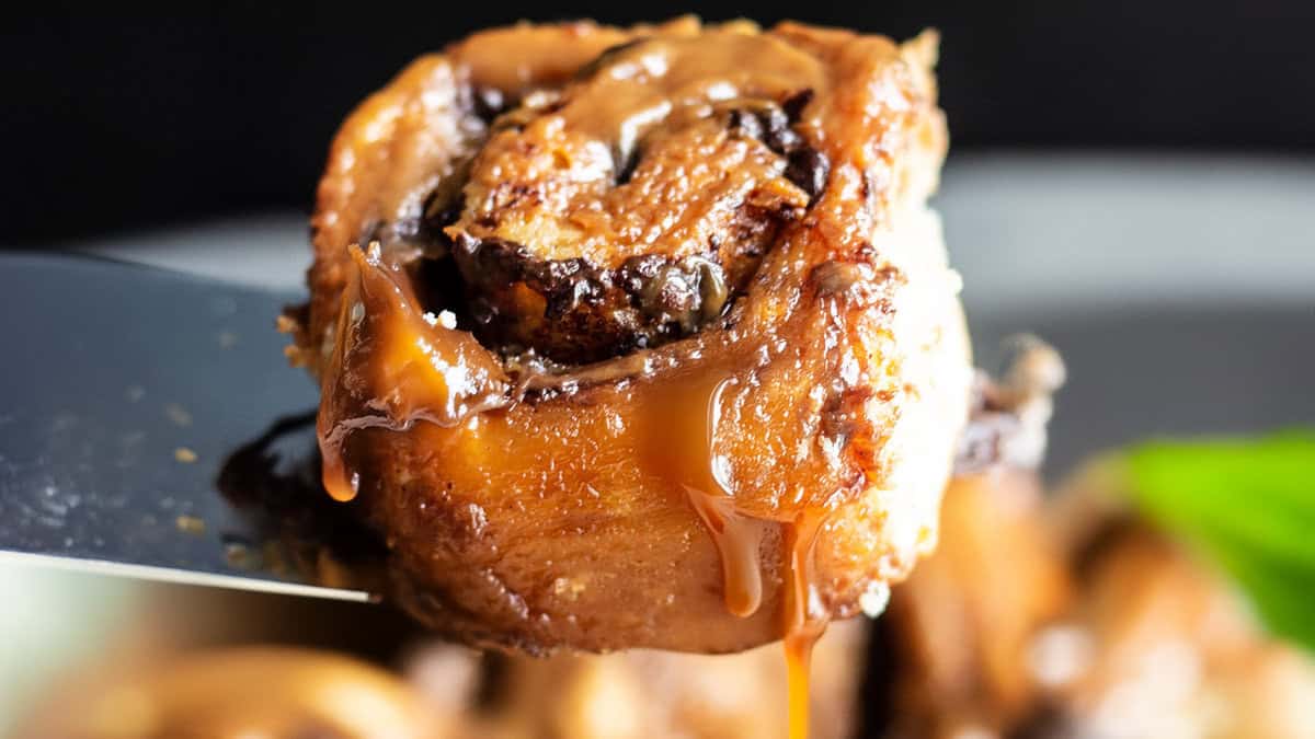 Close-up of a gooey caramel chocolate sticky bun being lifted by a spatula. The pastry is generously coated with caramel sauce, which drips down its sides, highlighting its rich, sticky texture. The background is blurred, similar to mouth-watering pancake recipes, emphasizing the focus on the cinnamon roll.