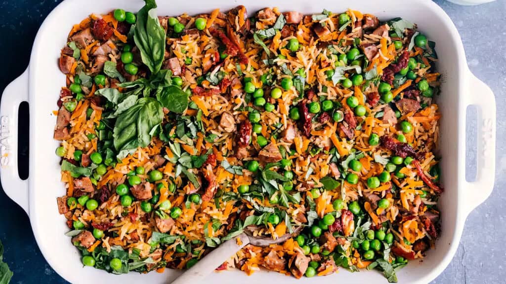 A white baking dish filled with a colorful mixed vegetable casserole, consisting of rice, diced chicken, peas, carrots, sun-dried tomatoes, and garnished with fresh basil leaves. A wooden serving spoon is placed inside the dish. Discover more delightful rice dish recipes to elevate your meals.