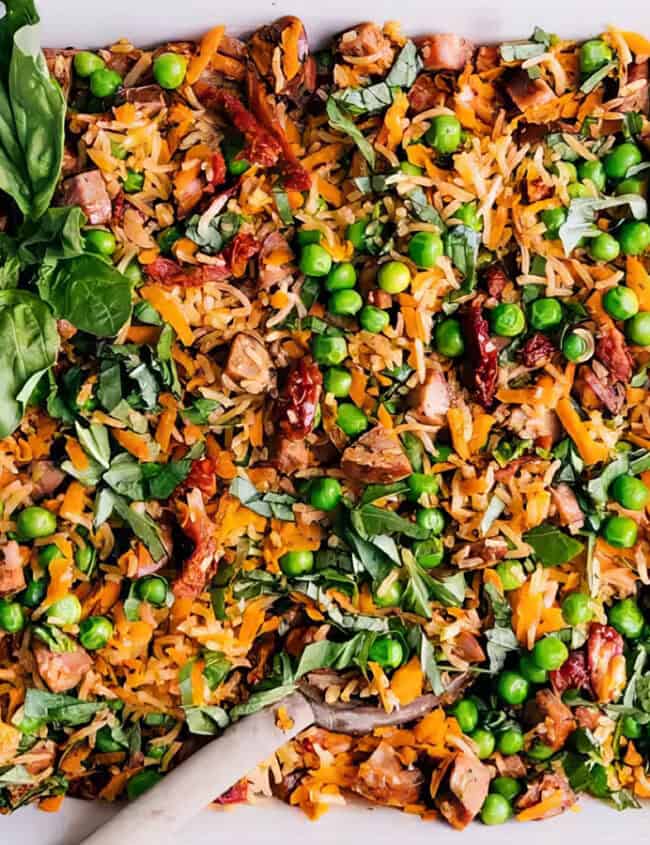 A white baking dish filled with a colorful mixed vegetable casserole, consisting of rice, diced chicken, peas, carrots, sun-dried tomatoes, and garnished with fresh basil leaves. A wooden serving spoon is placed inside the dish. Discover more delightful rice dish recipes to elevate your meals.