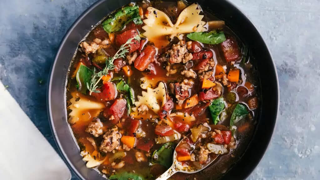 A hearty bowl of soup filled with farfalle pasta, ground meat, diced tomatoes, carrots, spinach, and fresh herbs served in a black bowl. A spoon is partially submerged in the flavorful, colorful broth. This comfort food warms your soul against a light blue surface in the background.