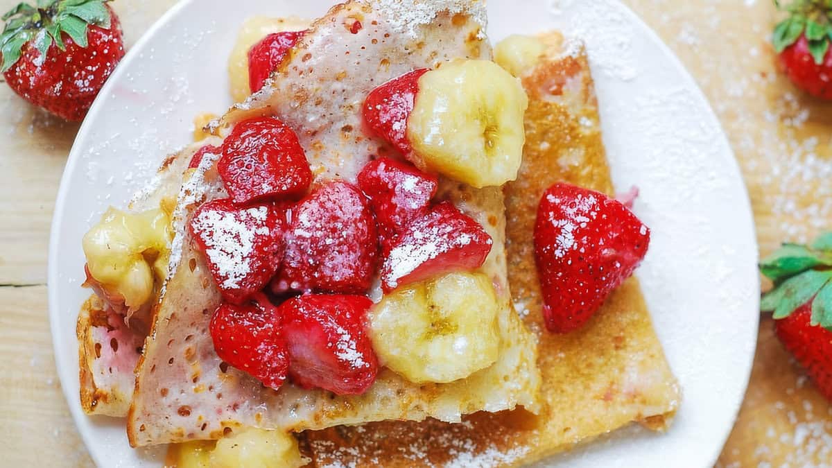 A close-up of a plate with crepes topped with powdered sugar, strawberries, and banana slices. The light golden brown crepes are drizzled with peanut butter, satisfying your sweet cravings. Fresh strawberries are scattered around the plate on a wooden surface, adding a touch of color to your delicious breakfast.