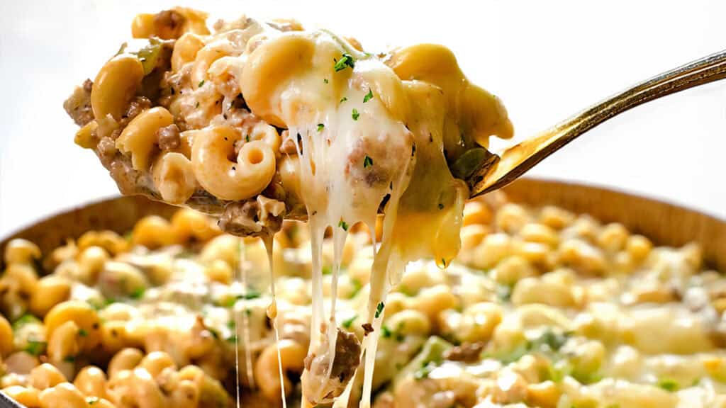 A close-up of a spoon lifting a cheesy portion of baked macaroni and beef from a dish, showcasing one of the delicious ground beef recipes. The pasta is covered in melted cheese and garnished with chopped herbs, with the remaining macaroni and cheese visible in the background.