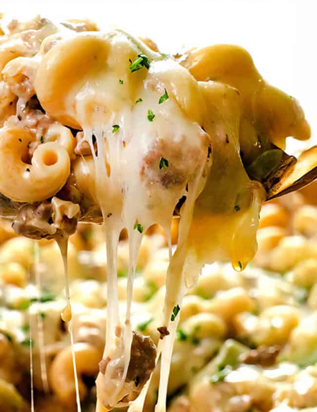 A close-up of a spoon lifting a cheesy portion of baked macaroni and beef from a dish, showcasing one of the delicious ground beef recipes. The pasta is covered in melted cheese and garnished with chopped herbs, with the remaining macaroni and cheese visible in the background.