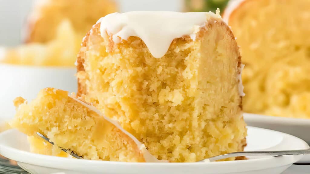 A close-up of a slice of pineapple-flavored Bundt cake on a white plate. The cake is moist and dense with chunks of pineapple throughout, topped with a dripping glaze. A small forkful of the cake is resting on the plate, perfect for those seeking delicious Bundt Cake recipes.