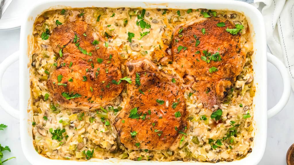 A white casserole dish filled with three browned pork chops resting on a creamy mixture of rice, mushrooms, and herbs. This Southern recipe is garnished with fresh chopped parsley.