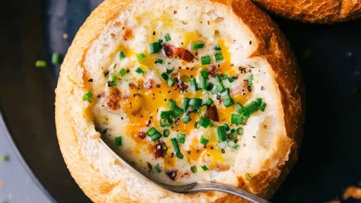 Close-up of a bread bowl filled with creamy soup, topped with chopped chives, bits of bacon, black pepper, and melted cheese. A spoon is partially dipped into the soup, highlighting the rich texture and garnishes—a true comfort food to warm your soul. Perfect for those looking for delicious soup recipes.