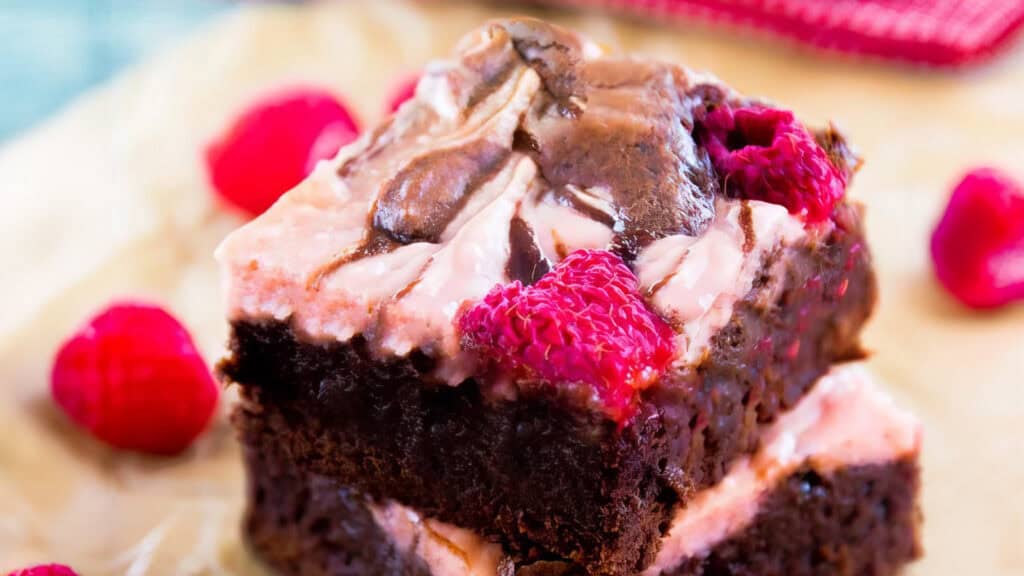 A close-up shot of a dessert with two stacked fudgy chocolate brownies, perfect for chocoholics, topped with a layer of raspberry cream cheese frosting and fresh raspberries. Some raspberries are scattered around the brownies on a light-colored surface.