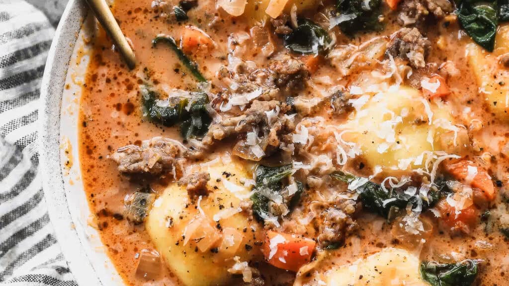 A hearty bowl of soup with ground beef, spinach, and pasta dumplings garnished with grated cheese. The soup appears thick and creamy with visible chunks of vegetables. A spoon rests in the bowl, and a striped cloth is partially visible underneath—truly a delightful addition to your ground beef recipes.