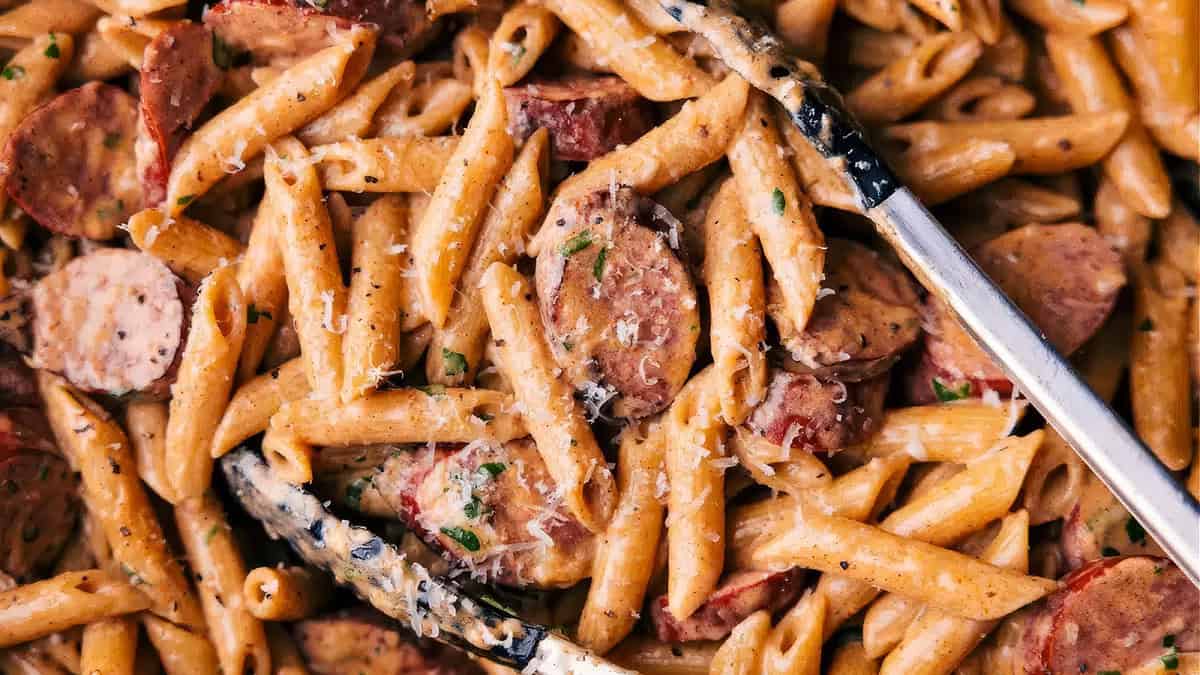 A close-up of creamy penne pasta mixed with slices of sausage, sprinkled with grated cheese, and garnished with finely chopped herbs. The dish, inspired by Alfredo recipes, is being served with a pair of tongs.