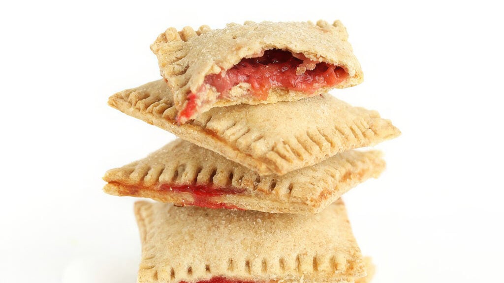 A stack of three square pastries, classic strawberry desserts, is shown against a white background. The top pastry has a bite taken out of it, revealing a red fruit filling inside. The pastries have a golden-brown crust and a crimped edge design.