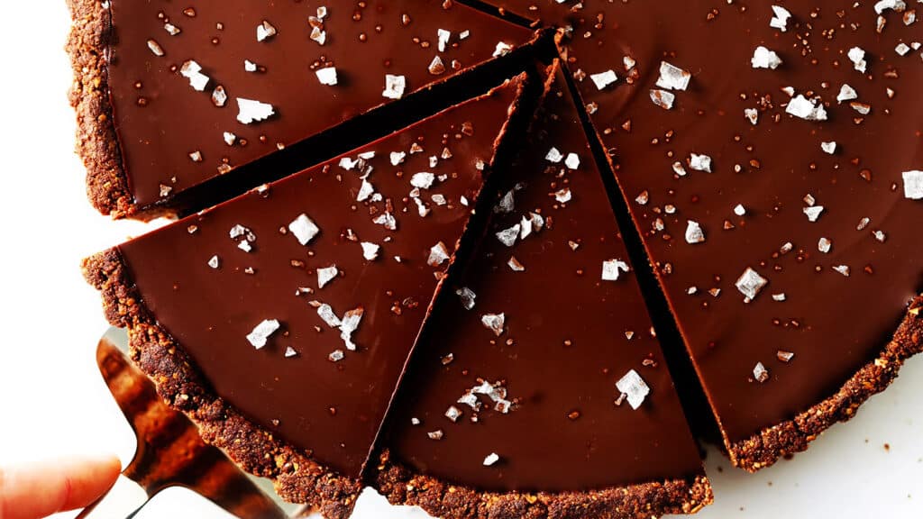 A rich chocolate tart sliced into six pieces. The smooth, glossy chocolate filling is topped with flakes of sea salt, and the tart has a dark, crumbly crust. A server is lifting one slice away from the tart, demonstrating one of the finest chocolate recipes. The background is white and minimalistic.