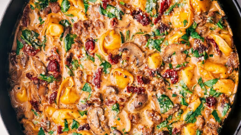 A close-up of a creamy tortellini dish featuring cheese-filled pasta, sliced mushrooms, sun-dried tomatoes, ground meat, and fresh spinach. The dish is garnished with chopped herbs and has a rich, savory sauce.