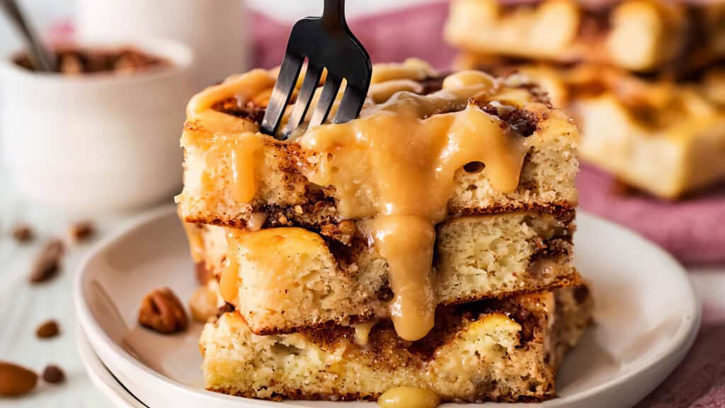 A close-up of a decadent cinnamon roll cake slice stacked in two layers on a white plate, reminiscent of pancake recipes. It is generously drizzled with caramel icing, with a fork piercing the top slice. The background shows a blurry setting with more cake pieces and a bowl with toppings.