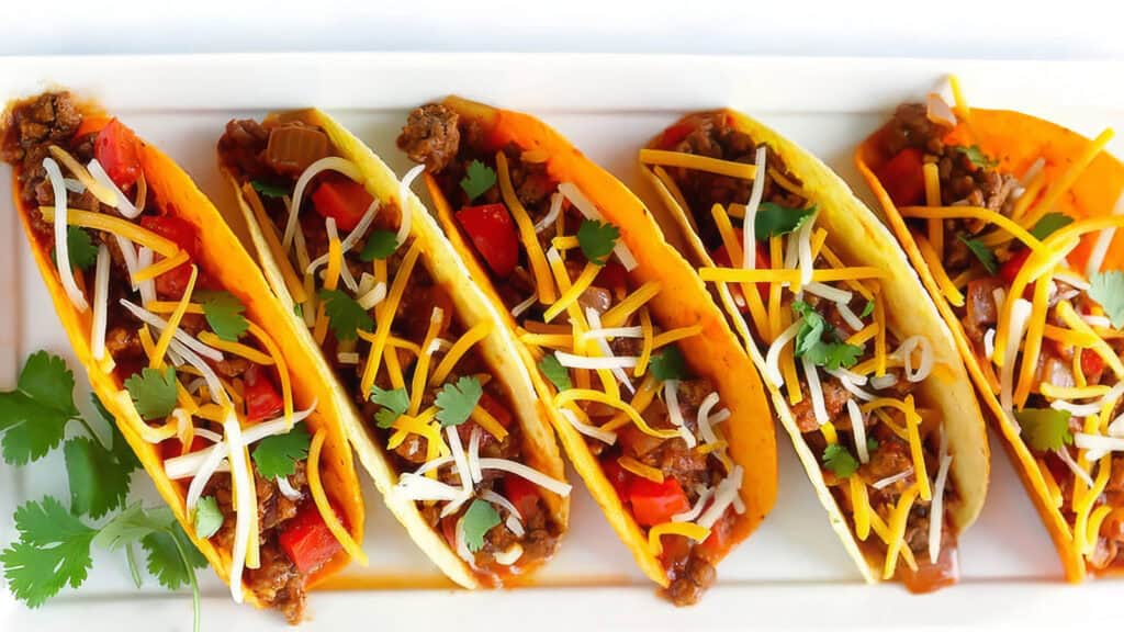 A row of five hard shell tacos filled with seasoned ground beef, diced tomatoes, red bell peppers, shredded cheese, and garnished with cilantro—an impeccable addition to your favorite ground beef recipes—all arranged neatly on a white rectangular plate.