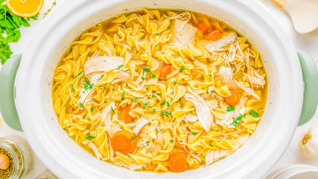 A slow cooker filled with chicken noodle soup, featuring wide egg noodles, shredded chicken, sliced carrots, and garnished with fresh herbs. The edges of ingredients like garlic, seasoning, and lemon are partially visible around the pot—a perfect addition to your slow cooker recipes collection.