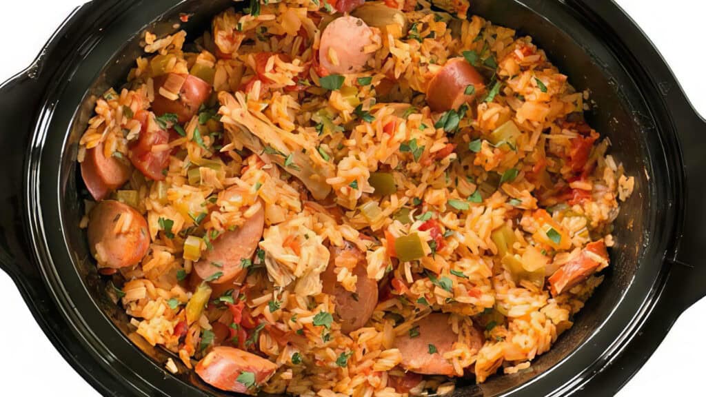 A slow cooker filled with a colorful rice dish. The dish contains sliced sausages, diced green peppers, and chunks of tomatoes, all mixed with seasoned rice. Garnished with chopped parsley, this hearty and flavorful creation is one of the best slow cooker recipes you can enjoy.