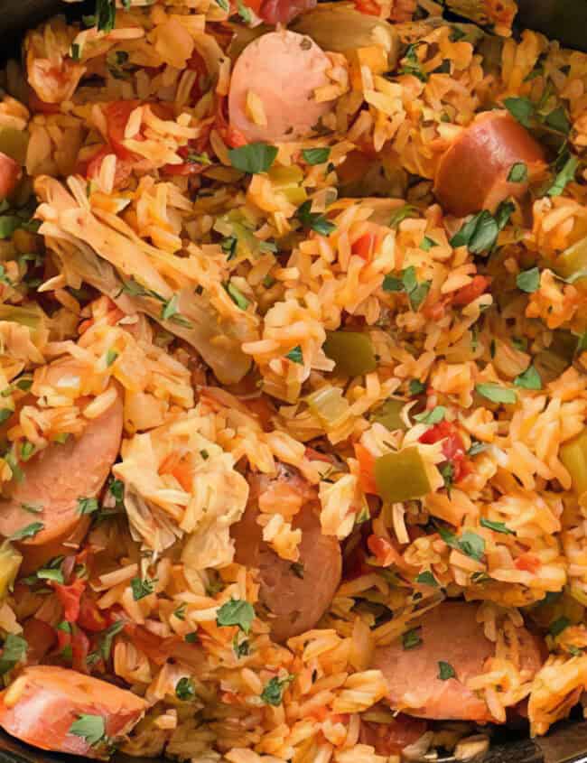 A slow cooker filled with a colorful rice dish. The dish contains sliced sausages, diced green peppers, and chunks of tomatoes, all mixed with seasoned rice. Garnished with chopped parsley, this hearty and flavorful creation is one of the best slow cooker recipes you can enjoy.