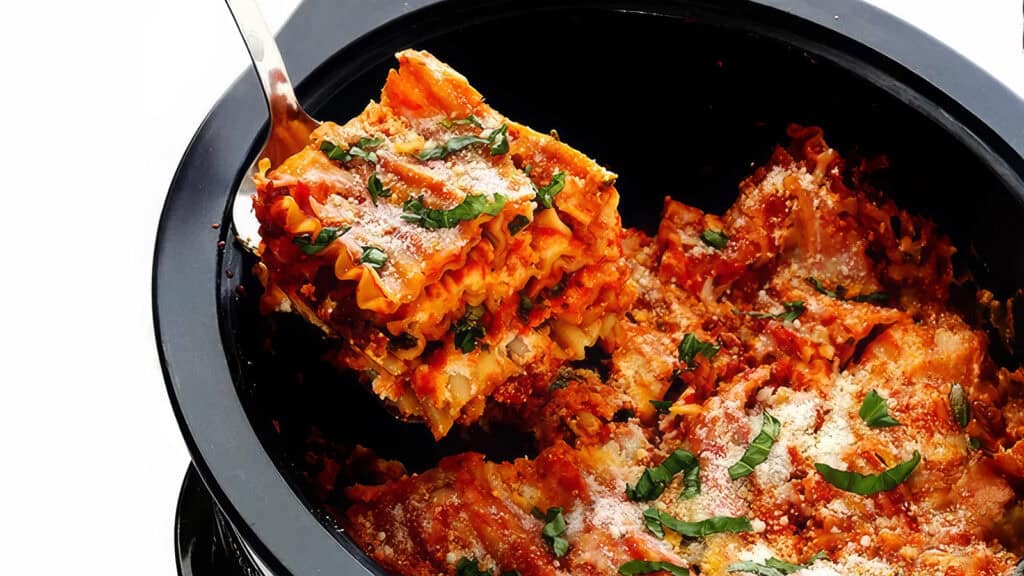 A close-up of a fork lifting a piece of lasagna out of a black slow cooker. The lasagna is layered with pasta, red sauce, and melted cheese, and garnished with fresh basil and grated Parmesan cheese. This hearty and well-cooked dish showcases the best of slow cooker recipes.