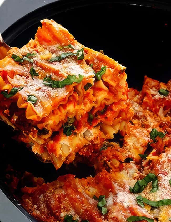 A close-up of a fork lifting a piece of lasagna out of a black slow cooker. The lasagna is layered with pasta, red sauce, and melted cheese, and garnished with fresh basil and grated Parmesan cheese. This hearty and well-cooked dish showcases the best of slow cooker recipes.