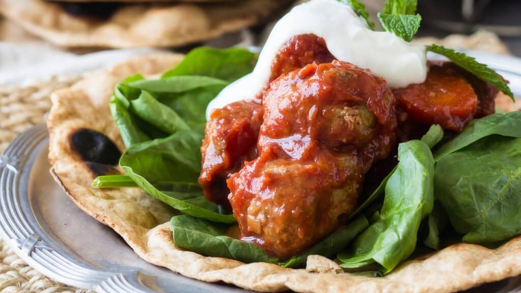 A close-up of a dish featuring a piece of flatbread topped with fresh spinach, tomato-based sauce, chunks of slow cooker recipe meat and vegetables, and a dollop of white creamy sauce. A sprig of mint garnishes the dish, which is served on a rustic metal plate.