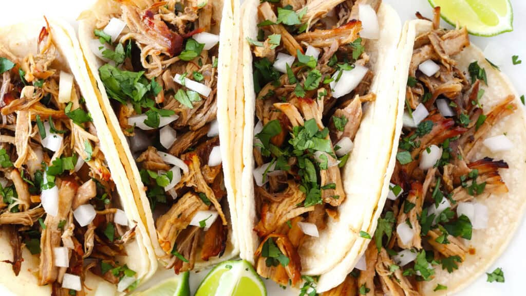 Three soft tacos filled with shredded meat, chopped onions, and cilantro are arranged in a row. Lime wedges are placed around the tacos, adding a fresh touch to this vibrant, appetizing meal inspired by slow cooker recipes.