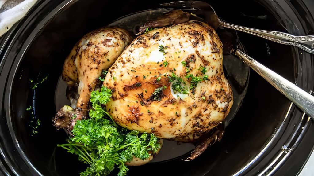 A cooked whole chicken, seasoned with herbs and garnished with fresh parsley, rests in a black slow cooker. Two metal utensils, a fork and a spoon, are placed inside the cooker alongside the chicken—an enticing addition to your collection of slow cooker recipes.