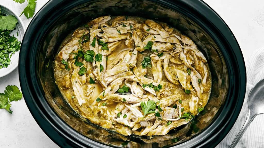 A slow cooker filled with shredded chicken in a green sauce, garnished with cilantro. This delightful dinner showcases one of the best slow cooker recipes, ready to be served with fresh cilantro visible in a small dish to the side.