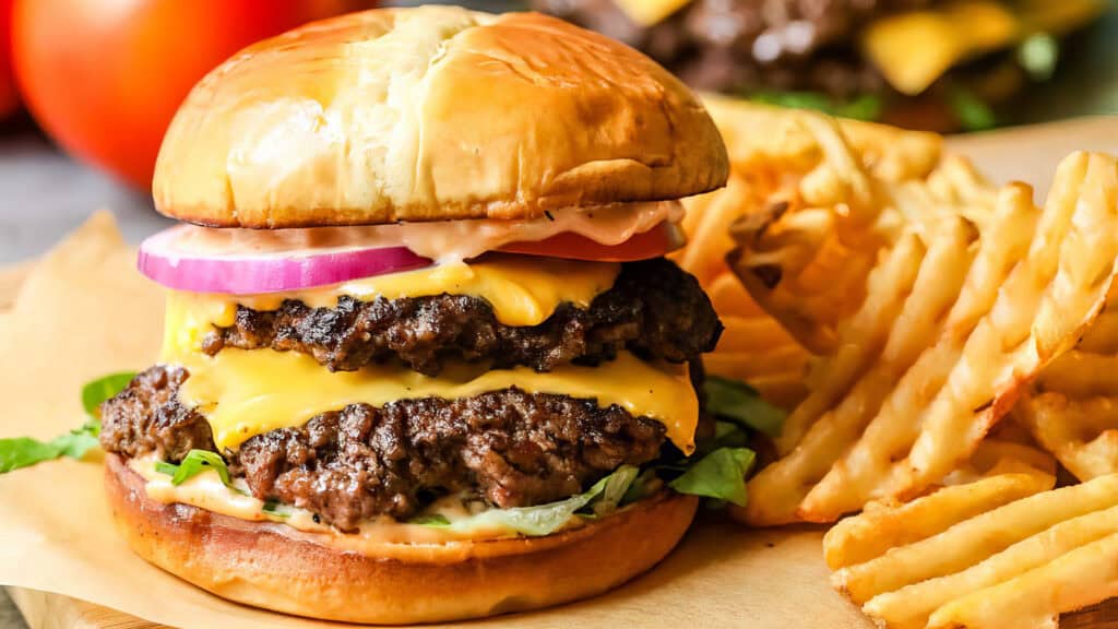 A juicy double cheeseburger with melted cheese, lettuce, tomato, and red onion slices is stacked high on a toasted bun. This delight from our ground beef recipes is served with a side of crispy waffle fries on a parchment-lined tray.