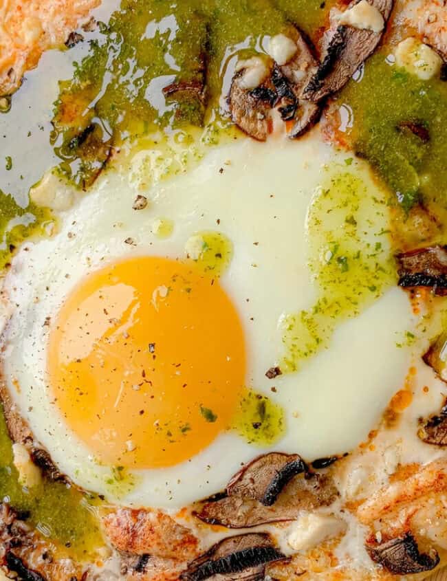 A close-up of a pizza topped with a sunny-side-up egg in the center, surrounded by mushrooms, melted cheese, and drizzled with green pesto sauce. Perfect for breakfast, lunch, and dinner, the pizza crust is crisp and golden brown.